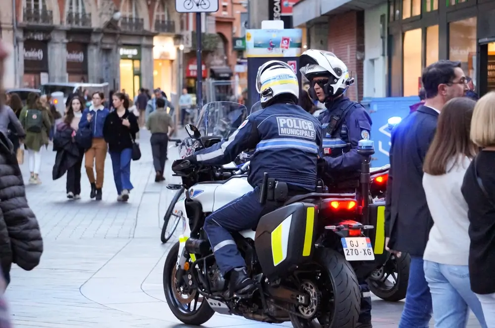 Policiers municipaux en patrouille à moto dans une rue piétonne, portant leur tenue et équipement de police.