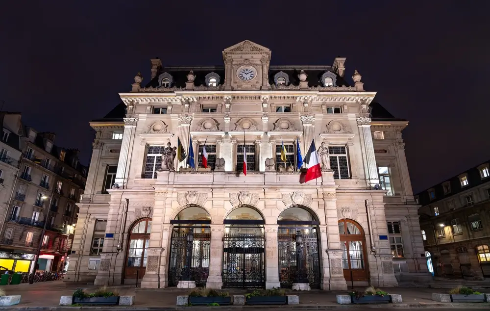 Façade illuminée d'une mairie de Paris, bâtiment administratif orné de drapeaux français devant l'entrée principale.